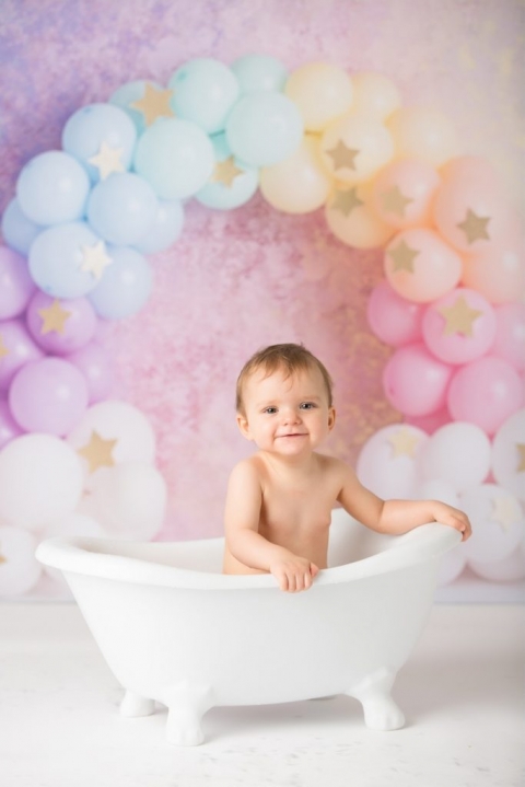 Baby enjoying a splash photo session after a cake smash session in a rolled top bath, with rainbow balloons and pastel garland